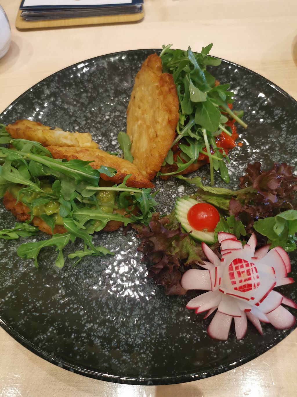 A dark gray plate on a light wood surface. On the plate, bottom right, is a small red raddish, which has been carved to resemble a flower. Top center and left are two potato cakes. One is sitting on a small pile of green guacomole, the other one on a pile of red tomato relish. There are several green salad leafs, a small red cherry tomato, and a slice of cucumber visible, too.