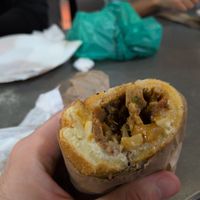 A Cheesesteak sandwich. White bread with seitan and onions in the middle. Behind the sandwich are a paper plate and green plastic bag resting on a table. at LUHV Vegan Deli in Philadelphia