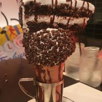 BLACK ‘N WHITE CAKESHAKE CHOCOLATE SHAKE: Vanilla frosted rim with mini chocolate chips topped with a black ‘n white cake slice, whipped cream & chocolate drizzle (AVAILABLE VE at Venetian Hotel - Black Tap in Las Vegas