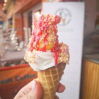 Photo of a hand holding an ice cream sugar cone, filled with vanilla whippy ice cream topped with lemon sorbet and covered with rainbow sprinkles and raspberry sauce at Harbourside Flavours in Bridlington
