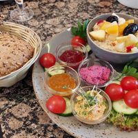 vegan breakfast plate: 3 rolls (left side), 4 spreads (2 jams, beetroot & bell pepper), warm oat milk porridge with fruits; some tomatoes & sliced cucumber at Café Fabrice in Schluechtern