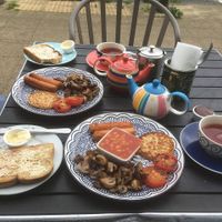 Outdoor table loaded with colourful teapots and cups with a vegan cooked breakfast of sausages, beans, potato cake, tomatoes and mushrooms on a blue and white plate. Yum!  at Driftwood Cafe in Wallasey