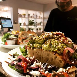 The Tuscan Avocado Toast has grilled foccacia with roasted peppers, marninated mushrooms, red onoin, greens, pistacios and goat cheese. We can omit the cheese to make it vegan. at Brunch in Conshohocken