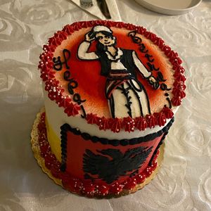 Top of cake features a woman dressed in men’s traditional Albanian costume, with happy birthday intentionally spelled wrong. Side of cake features Albanian double-headed eagle.  at Cakerino in Troy