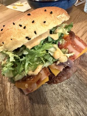 A close-up of a juicy plant-based burger with lettuce, tomato, onions, cheese, sauce, and a seeded bun, served on a wooden board. Another photo shows the fully vegan menu.  at Tabi Loo in Leuven