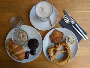 Latte with oat milk,left plate with croissant, Bircher muesli,chocolate muffin, jam and chocolate spread, right plate with pretzel, Bavarian meatloaf ("Leberkäse") and basil sprea at KOOS Bowls & Bar in Munich