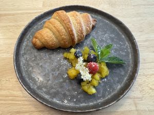 Frisches Croissant mit Fruchttatar aus Mango, garniert mit Beeren, Minze und Holunderblüten – liebevoll angerichtet auf einem dunklen Teller.  at Ti's Café in Minheim