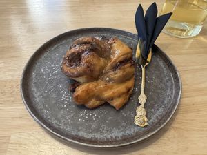 Ein goldbraun gebackener Dattel-Kardamom-Zopf liegt auf einem rustikalen, dunklen Keramikteller. Der Teig ist glänzend karamellisiert und kunstvoll verdreht.  at Ti's Café in Minheim