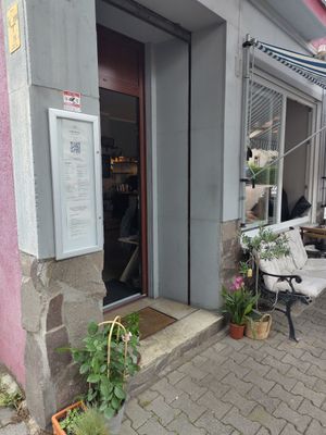 Eingangsbereich mit Pflanzen. Auf dem Bürger*innensteig vor dem Laden stehen Tische und Stühle und eine bequeme Bank. at Nunu Mannheim in Mannheim