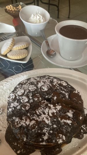 3 pancakes con topping di cioccolata gusto oreo, gocce di cioccolato al latte vegan e zucchero a velo. Dietro cioccolata calda alla nocciola, panna montata veg, biscottini.  at Yogurteria Sui Generis in Salerno