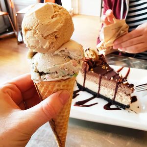1.5 scoops of ice cream (birthday cake and pumpkin spice) in a waffle cone, with a slice of chocolate peanut butter cheesecake in the background. at Cosmic Treats in Toronto
