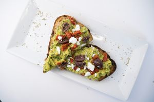 Mediterranean Avocado Toast made from Avocado, sun dried tomato, vegan feta, spinach, vegan basil aioli (contains nuts), olives, olive oil, Italian seasoning, s+p on olive bread at Glowing Plant Based Eatery in Los Angeles