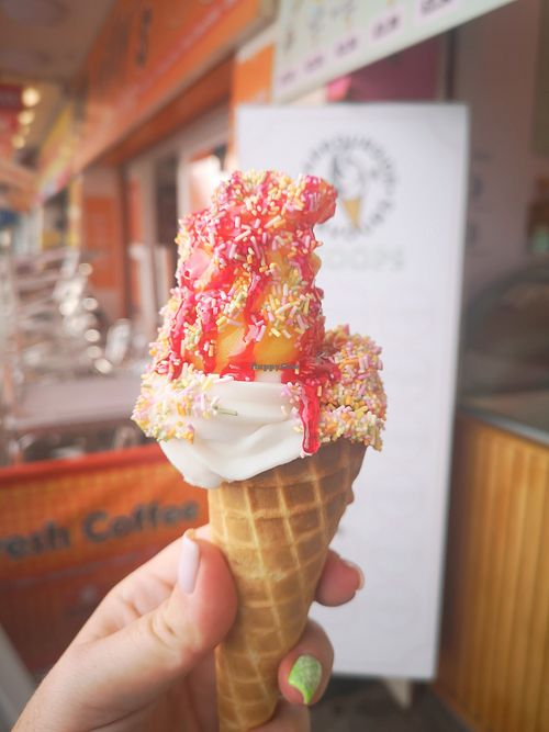 Photo of a hand holding an ice cream sugar cone, filled with vanilla whippy ice cream topped with lemon sorbet and covered with rainbow sprinkles and raspberry sauce at Harbourside Flavours in Bridlington