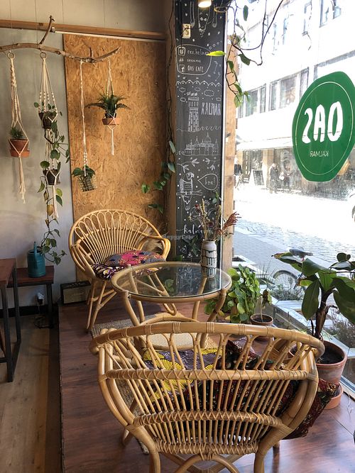 Picture of a glass table and wicker chairs in front of the café’s full-length storefront window. There are plants hanging from a branch in the background, and other plants aroun at OAS Cafe in Kalmar